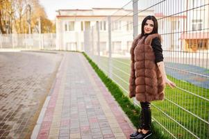 photo de mode en plein air d'une magnifique femme sensuelle aux cheveux noirs dans des vêtements élégants et un luxueux manteau de fourrure sans manches contre une clôture de fer à la ville d'automne.