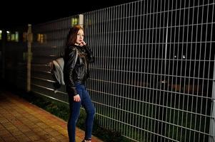 portrait de nuit d'une fille modèle portant un jean et une veste en cuir contre une clôture en fer. photo