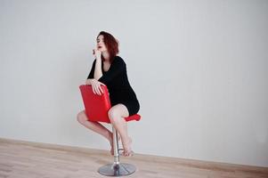 fille aux cheveux rouges sur une tunique de robe noire assise sur une chaise rouge contre un mur blanc dans une salle vide. photo