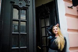 fille blonde à la mode en long manteau de cuir noir posé contre la porte en bois de la vieille maison. photo