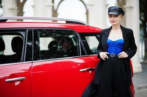 portrait d'une belle mannequin blonde sexy en casquette et en corset noir et bleu, avec un maquillage lumineux près d'une voiture de ville rouge. photo