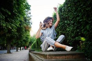 portrait d'une superbe jeune femme en combinaison rayée assise dans le parc et écoutant de la musique avec ses écouteurs. photo