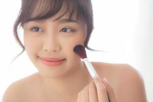 portrait de beauté jeune femme asiatique sourire avec le visage regardant le miroir se maquiller avec la joue de la brosse dans la chambre, belle fille tenant le fard à joues, les soins de la peau et le concept cosmétique. photo