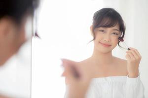 portrait de beauté jeune femme asiatique souriante avec un miroir à la recherche de visage appliquant du maquillage avec une joue de brosse dans la chambre, belle fille tenant un fard à joues, des soins de la peau et un concept de mode de table cosmétique. photo