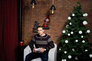 portrait en studio d'un homme avec un livre assis sur une chaise contre l'arbre de noël avec des décorations. photo