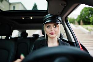 portrait d'une belle mannequin blonde sexy en casquette et en noir avec un maquillage lumineux s'asseoir et conduire une voiture de ville rouge. photo