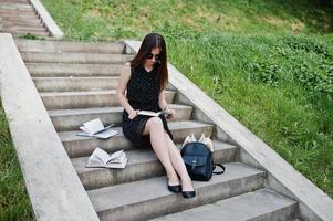 portrait d'une belle femme en robe noire à pois et lunettes de soleil assis sur les escaliers avec des livres et sac à dos dans le parc. photo