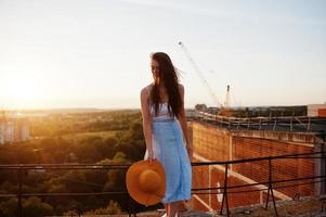 portrait d'une jolie fille en vêtements décontractés posant sur le toit avec son chapeau à la main au coucher du soleil. photo