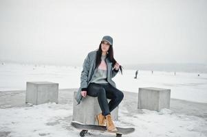 fille brune élégante en casquette grise, style de rue décontracté avec planche à roulettes le jour de l'hiver. photo