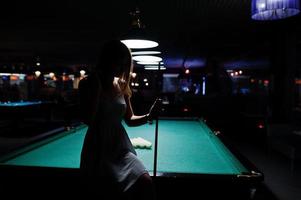 portrait d'une jolie jeune femme en robe assise sur la table de billard et tenant une queue. photo