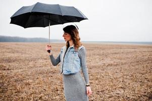 portrait de jeune fille bouclée brune en veste jeans avec parapluie noir sur le terrain. photo