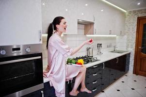 portrait d'une belle jeune femme en robe rose posant avec une tomate dans sa cuisine. photo