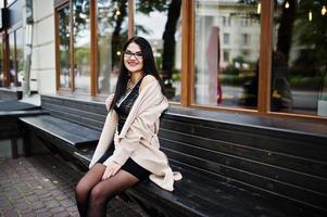 femme sexy cheveux noirs à lunettes et manteau assis sur un banc. photo