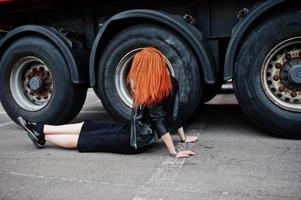 fille élégante aux cheveux rouges en noir, assise contre de grandes roues de camion. photo