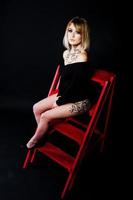 portrait en studio d'une fille blonde avec à l'origine maquillage sur le cou et tatouage sur la cuisse, porter sur une robe noire sur fond sombre, assis sur une échelle rouge. photo