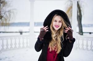 élégance fille blonde en manteau de fourrure et robe de soirée rouge posée au jour de neige d'hiver. photo