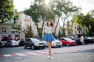 fille bouclée et élégante porte une jupe en jean bleu, un chemisier et des lunettes posées dans la rue de la ville avec une tasse de café. photo