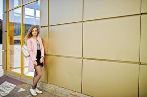 jeune fille blonde en jupe noire et manteau rose posé contre un mur de texture dorée. photo