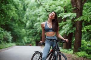 beau temps aujourd'hui. Cycliste féminine debout avec vélo sur route goudronnée dans la forêt pendant la journée photo