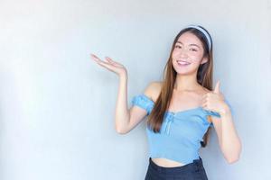 jeune belle femme asiatique dans une chemise bleue agit le pouce vers le haut comme un bon symbole et une autre main montre comme présentant quelque chose sur le fond blanc. photo