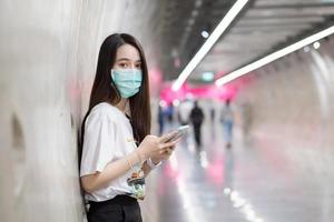 une jeune femme asiatique se tient dans le tunnel du métro et porte un masque facial comme nouveau mode de vie normal et de soins de santé tout en tenant un smartphone dans sa main. photo