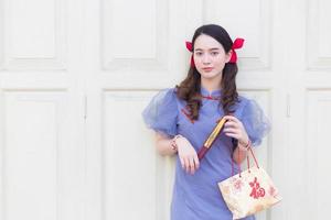 une femme asiatique en robe qipao chinoise bleu-gris tient le sac en tissu qui a projeté un mot qui signifie heureux en chinois et se tient sur une porte en bois blanc comme arrière-plan. photo