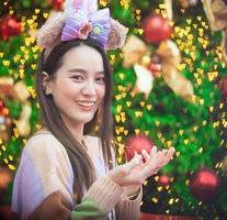 belle femme asiatique dans un chandail coloré se tient joyeusement. dans sa main, il tenait une lumière devant l'arbre de Noël. avec bokeh en forme de coeur comme arrière-plan sur le thème de noël et du nouvel an photo