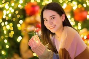 belle femme asiatique dans un chandail coloré se tient joyeusement. dans sa main, il tenait une lumière devant l'arbre de Noël. avec bokeh en arrière-plan sur le thème des fêtes de noël et du nouvel an photo