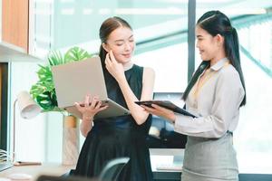 deux jeunes femmes d'affaires discutant d'un projet et d'un rapport sur un ordinateur portable et une tablette numérique tout en se tenant debout et en analysant des données sur les statistiques et des informations de remue-méninges dans un bureau moderne photo