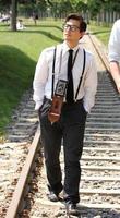jeune homme portant des lunettes et des bretelles marchant le long d'une voie ferrée avec un appareil photo vintage autour du cou