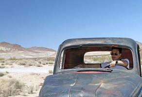 jeune homme portant des lunettes de soleil assis dans une épave de voiture abandonnée dans la vallée de la mort photo