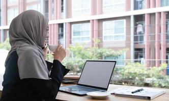 heureuse belle travailleuse musulmane tenant une tasse de café chaud assise au bureau et utilisant un téléphone portable en regardant les nouvelles en ligne se détendre. photo