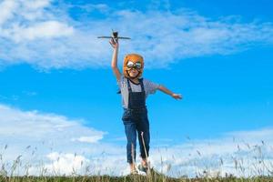 jolie petite fille qui traverse la prairie par une journée ensoleillée avec un avion jouet à la main. enfant heureux jouant avec un avion en carton sur fond bleu ciel d'été. concept d'imagination de rêve d'enfance.