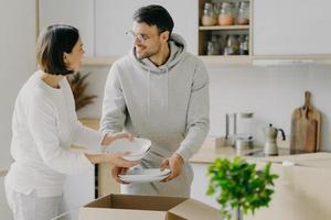 une femme au foyer ravie et son mari déballent des boîtes en carton avec de la nouvelle vaisselle, transportent des assiettes, emménagent dans un nouvel appartement, se regardent avec joie, posent contre l'intérieur de la cuisine. notion de déménagement