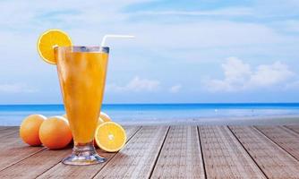 jus d'orange dans un verre transparent avec des glaçons et du jus d'orange à l'embouchure du verre. boissons fraîches posées sur le balcon en bois au bord de la mer en milieu de journée. rendu 3d photo