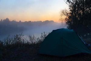 tente touristique verte au bord de la rivière au lever du soleil, avec brouillard d'automne matinal sur l'eau. paysage touristique en plein air. photo