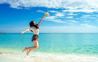 heureuse jeune femme à la mode de style décontracté et chapeau de paille sautant sur la plage de sable. détendez-vous et profitez de vacances sur une plage paradisiaque tropicale avec ciel bleu et nuages. fille en vacances d'été. ambiance estivale. photo