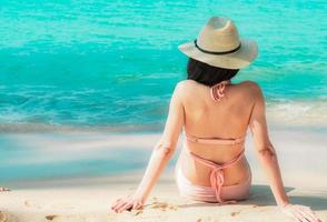 vue arrière d'une jeune femme asiatique heureuse en maillot de bain rose et chapeau de paille se détendre et profiter des vacances à la plage paradisiaque tropicale au coucher du soleil. fille à la mode des vacances d'été. modèle sexy de beauté. ambiance estivale. photo