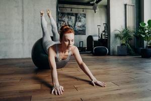 Dame mince athlétique aux cheveux rouges effectuant des exercices d'étirement sur un grand fitball argenté pendant l'entraînement de pilates photo