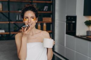 jeune femme caucasienne envoyant un message audio. fille applique des patchs oculaires anti-rides relaxants à la maison. photo