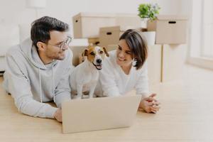 un joli couple juste marié allongé sur le sol, utilise un ordinateur portable, crée un projet de conception pour une nouvelle maison, leur chien préféré pose entre eux, une pile de boîtes en carton en arrière-plan. déménager dans une nouvelle maison photo