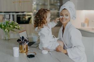 jolie petite fille aux cheveux bouclés regarde comment la mère fait les procédures cosmétiques applique des patchs de collagène sous les yeux pour le traitement de la peau porte des peignoirs pose près du bureau avec des produits de beauté. soins quotidiens photo