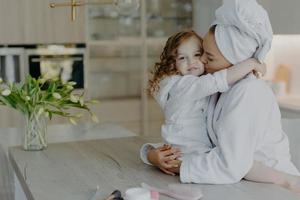 mère affectueuse portant une robe de chambre enveloppée d'une serviette de bain sur la tête embrasse et embrasse avec amour sa petite fille pose ensemble à la maison contre un intérieur confortable se sentir rafraîchi après avoir pris une douche photo