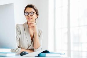 charmante employée de bureau garde la main sous le menton, regarde sur l'écran de l'ordinateur, porte des lunettes pour corriger la vision, pose dans son propre cabinet, prépare des informations pour une future réunion d'affaires photo