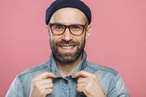 photo horizontale d'un homme barbu souriant et agréable portant des lunettes et une veste en jean, annonce de nouveaux vêtements à la mode, a une expression heureuse, isolé sur fond rose. Émotions positives
