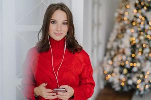 photo horizontale d'une jeune femme caucasienne qui aime la musique de la liste de lecture, écoute sa chanson préférée avec des écouteurs, vêtue d'un pull rouge tricoté, se dresse sur fond d'arbre de noël décoré