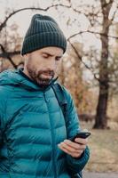 photo verticale d'un hipster concentré avec une barbe épaisse, porte un anorak et un chapeau, tient un téléphone portable, utilise une application spéciale pour trouver un itinéraire, un message avec un ami, respire de l'air frais. notion de mode de vie