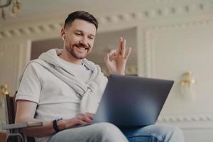 un homme barbu moderne fait un geste correct regarde avec plaisir l'écran de l'ordinateur portable porte des écouteurs a un appel vidéo porte un t-shirt jeans pull noué sur les épaules pose dans un appartement moderne. notion de technologie photo