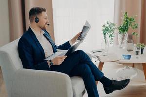 photo de côté d'un employé barbu d'une entreprise analyse les données regarde à travers des documents avec des graphiques examine le rapport financier vêtu de vêtements formels pose dans un appartement moderne utilise un casque d'ordinateur portable