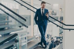 superbe entrepreneur en costume élégant formel debout sur l'escalier dans le hall du bureau avec smartphone photo